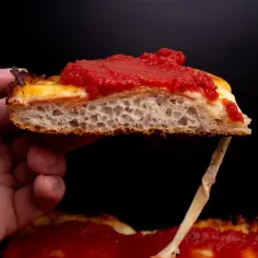 Kawałek puszystej detroit pizza z ciągnącym się serem i sosem pomidorowym, trzymany w dłoni.