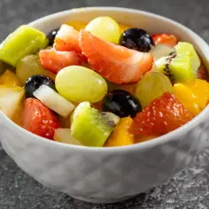 Kolorowa sałatka owocowa składniki: truskawki, kiwi, winogrona, mango i czarne porzeczki w białej miseczce.