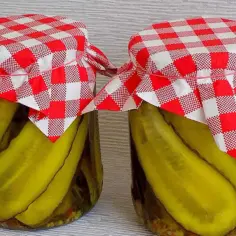 Dwa słoiki z ogórkami w curry ze skórką, ozdobione czerwono-białymi kratkowanymi chusteczkami.