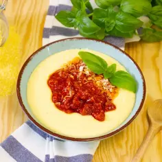 Aksamitna polenta z sosem pomidorowym i parmezanem, ozdobiona listkami bazylii. Obok słoik z mąką kukurydzianą.