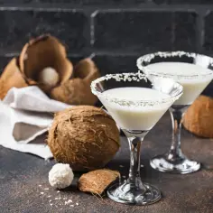 Dwa kieliszki z drinkiem Malibu z mlekiem, ozdobione wiórkami kokosowymi, obok świeżych kokosów.
