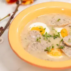 Biały barszcz przepis babci. Aromatyczna zupa z kiełbasą i jajkiem w pomarańczowej misce, ozdobiona zieleniną.