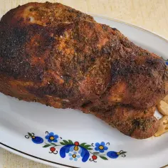 Rumiana łopatka wieprzowa, pieczona z ziołami, leży na talerzu ozdobionym ludowym wzorem. Obok kilka ząbków czosnku.