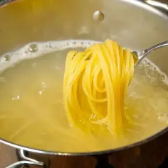 Z widelca zwisają gotujące się nitki makaronu spaghetti. Woda w garnku lekko bulgocze. To pyszne danie, które ma ok. 350 kcal w 100g surowego makaronu.
