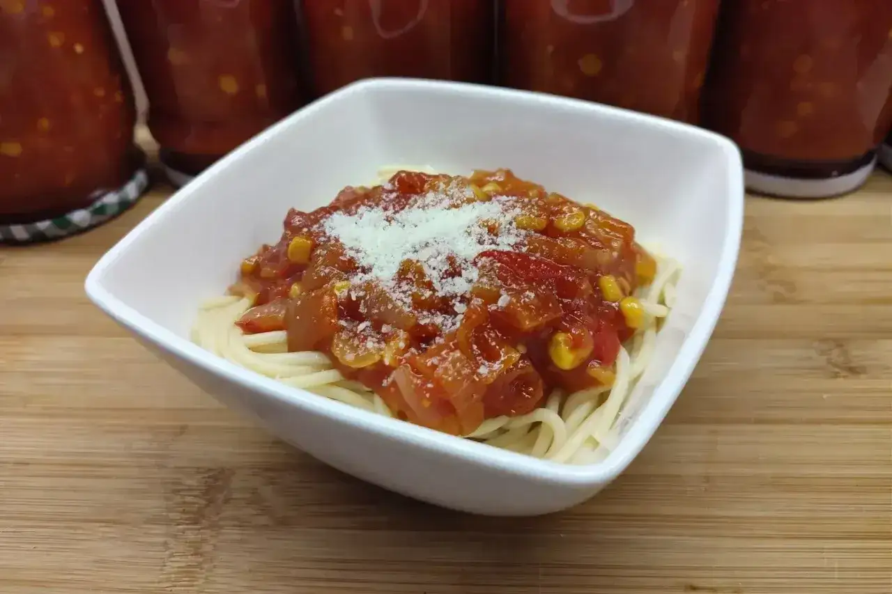 Jak zrobić sos do spaghetti? Na talerzu makaron z domowym sosem pomidorowym z kukurydzą i startym parmezanem. W tle słoiki z przetworami.