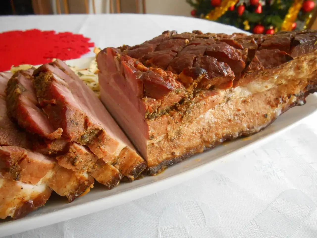 Pyszny boczek pieczony w rękawie, pokrojony w plastry, prezentuje się apetycznie na białej zastawie. W tle choinka.