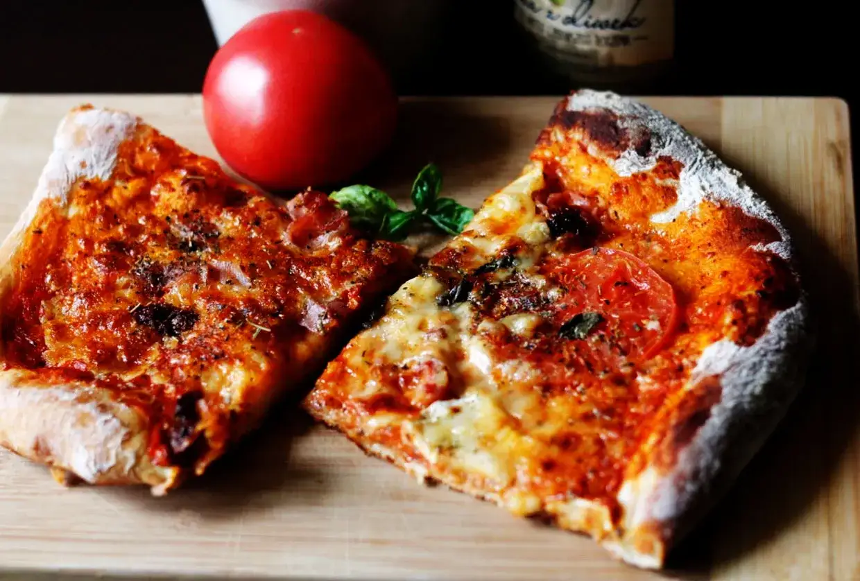 Dwie kromki pizzy na desce, obok pomidor. Idealny przepis na ciasto do pizzy, by stworzyć taki przysmak.