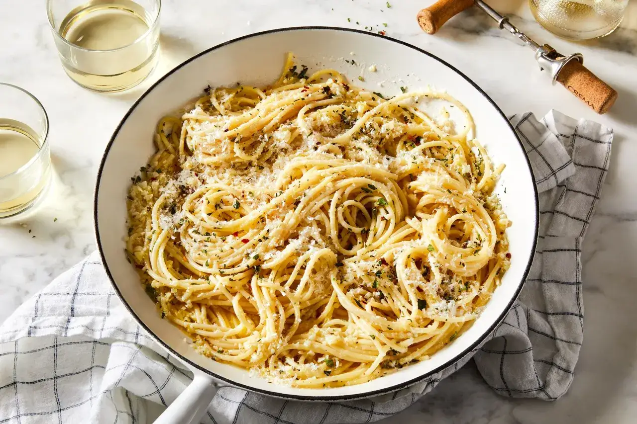 Pyszny sos do makaronu spaghetti z parmezanem i ziołami, podany w białej patelni, obok kieliszków białego wina.