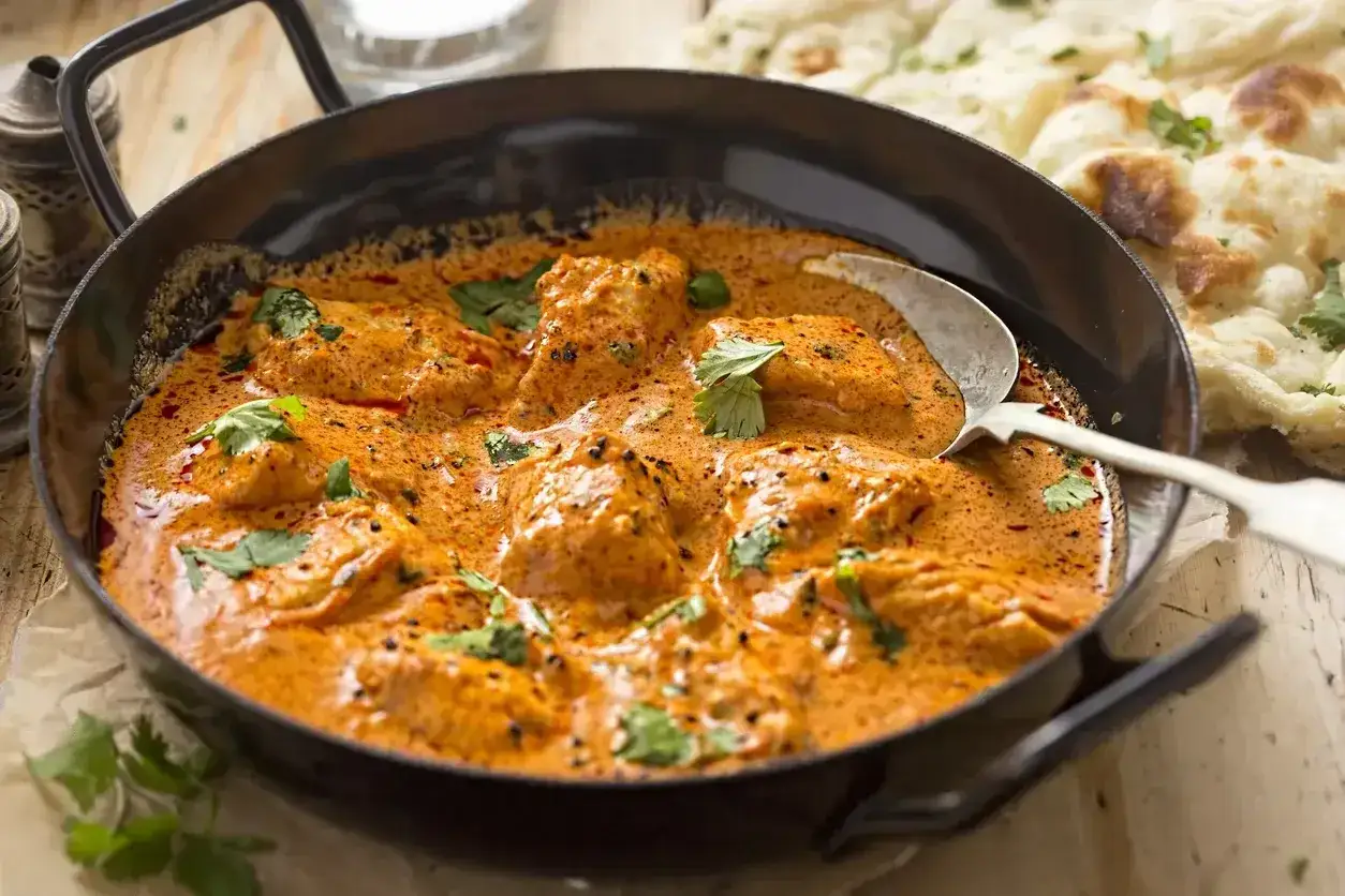 Aksamitny sos curry z kurczakiem, udekorowany świeżą kolendrą, podany w czarnej misie z łyżką. Obok widać kawałki chleba naan.