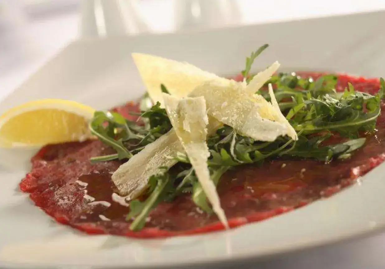 Eleganckie carpaccio wołowe z rukolą, płatkami parmezanu i plasterkiem cytryny.