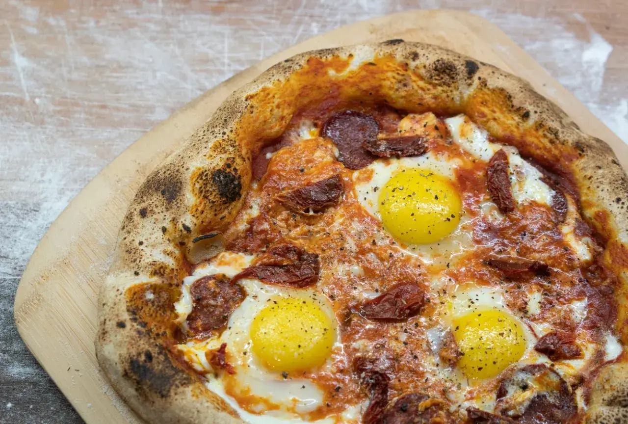 Pyszna pizza z jajkiem, salami i suszonymi pomidorami, z chrupiącym, lekko przypalonym brzegiem, na drewnianej desce.