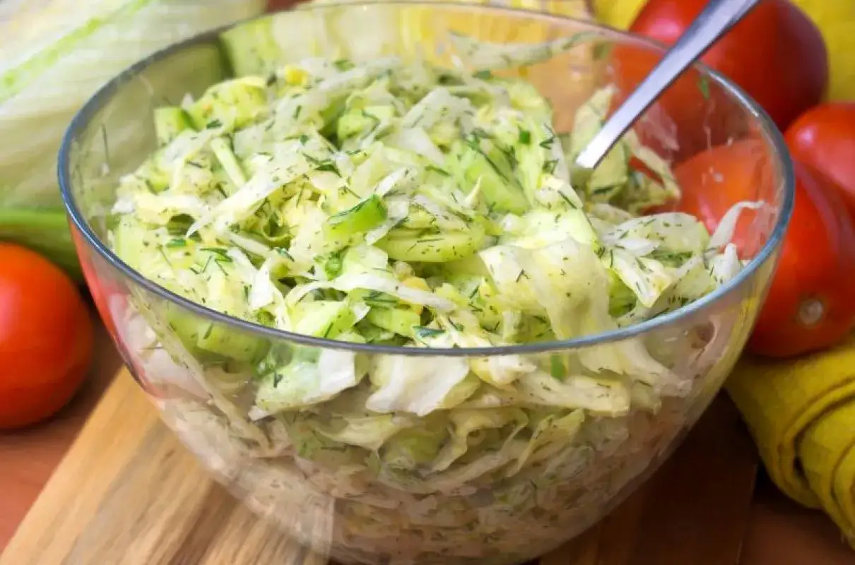 Chrupiąca surówka z sałaty lodowej, ogórka i koperku w szklanej misie, obok pomidory.