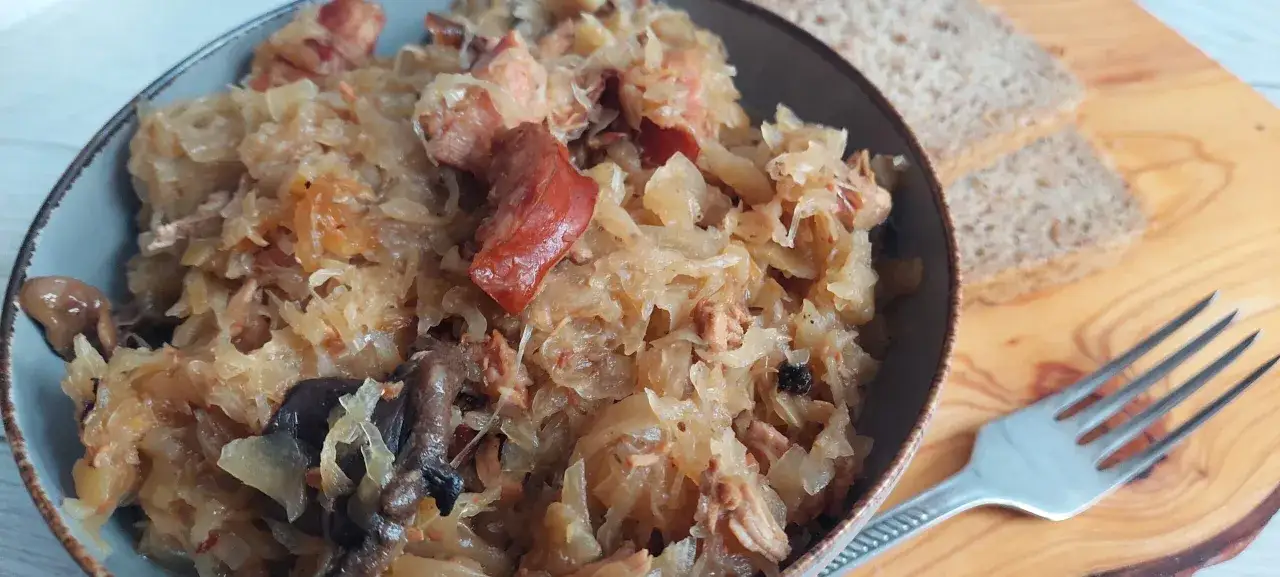 Misją jest znalezienie idealnego mięsa do bigosu. Na talerzu aromatyczny bigos z różnymi rodzajami mięsa, obok kromki chleba i widelca.