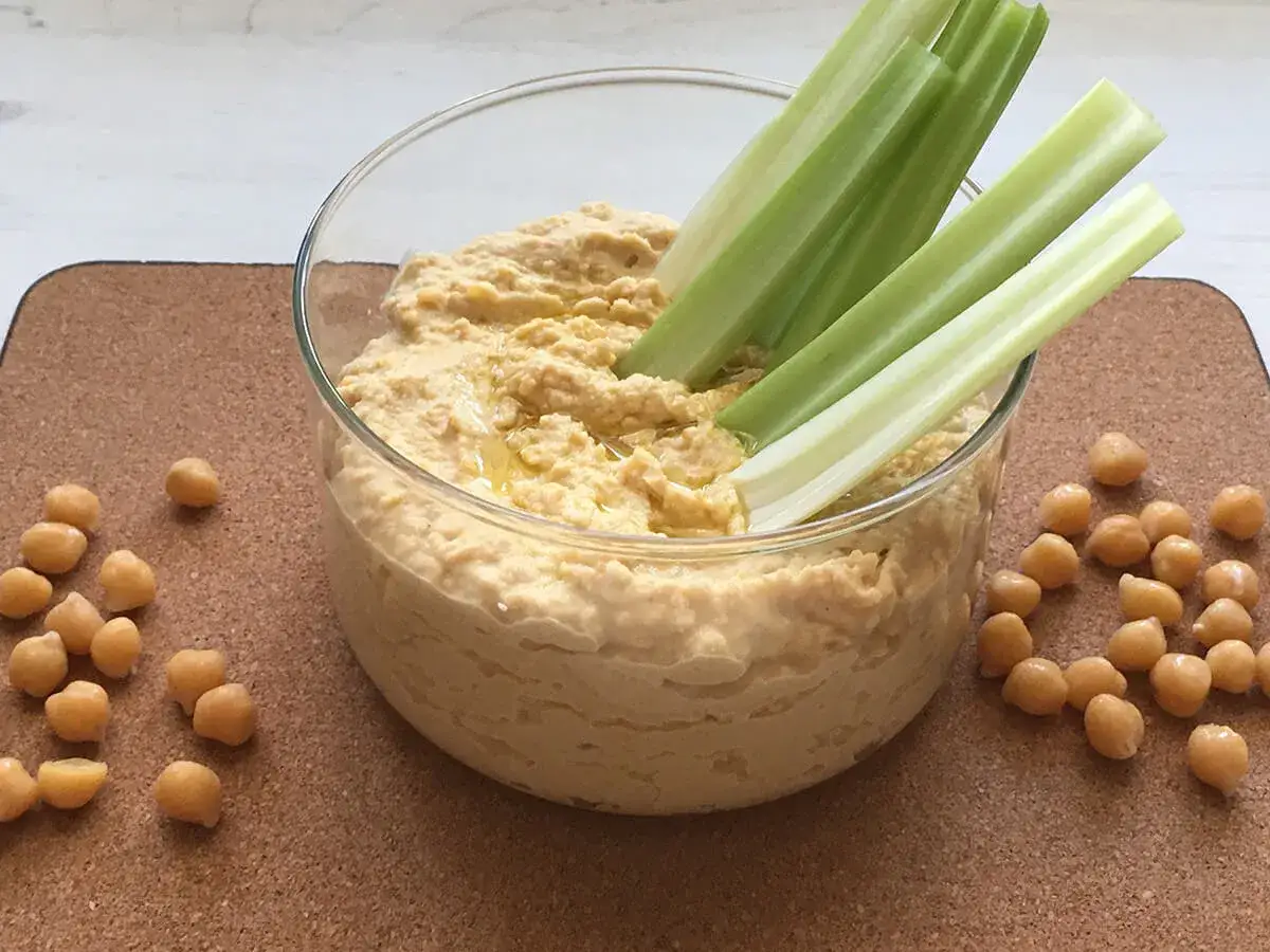 Domowy hummus przepis: kremowa pasta z ciecierzycy w szklanej misce, podana z łodygami selera i ziarnami ciecierzycy.