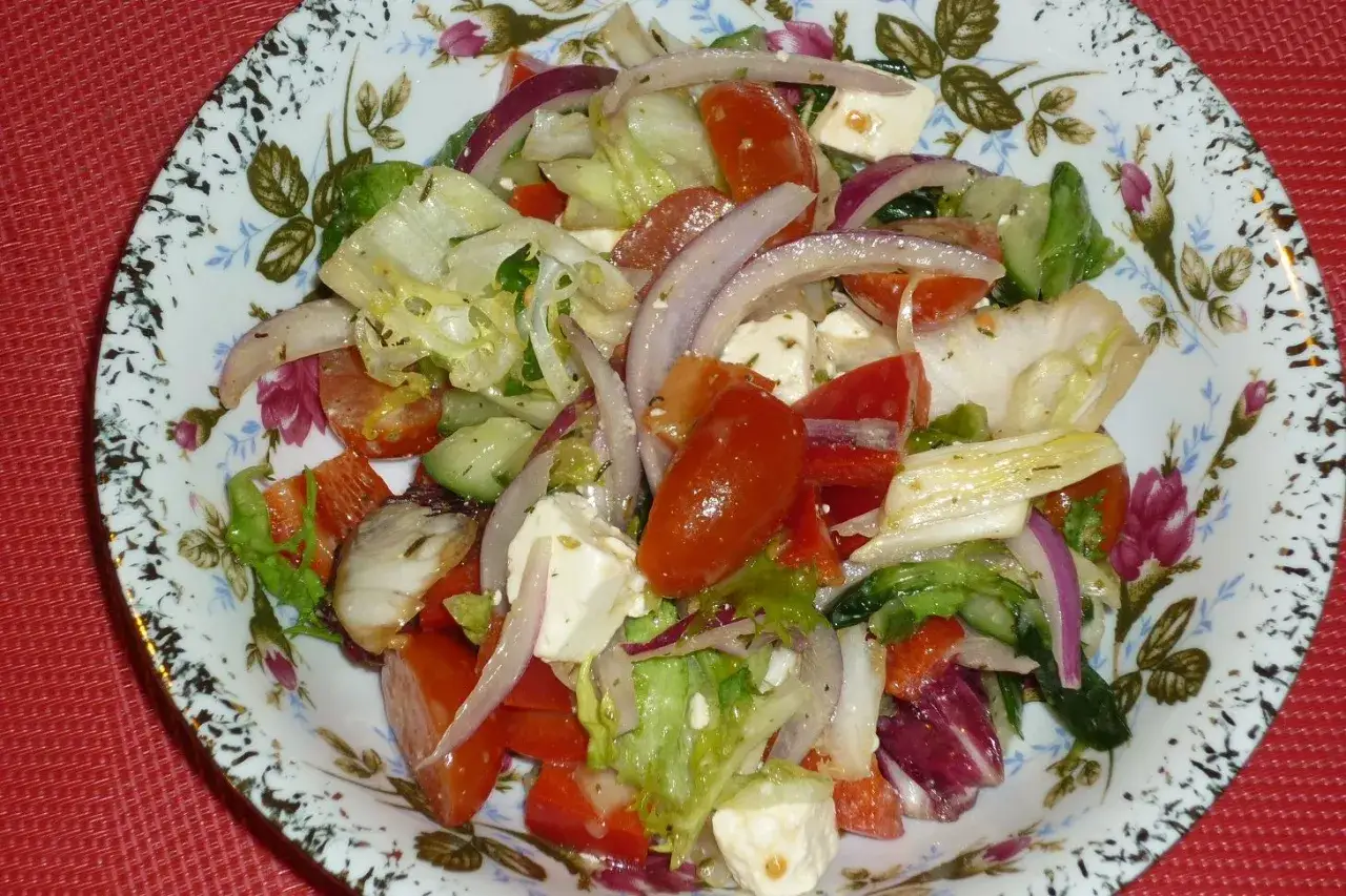 Kolorowa sałatka z mix sałat, pomidorów, ogórka, czerwonej cebuli i sera feta, podana na talerzu z kwiatowym wzorem.