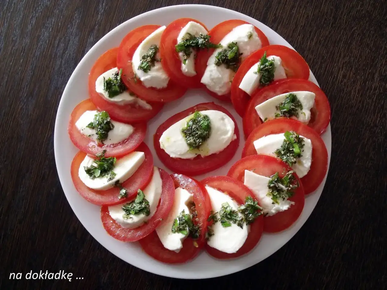 Kółka pomidorów z kawałkami mozzarelli i posiekaną bazylią, ułożone na talerzu.